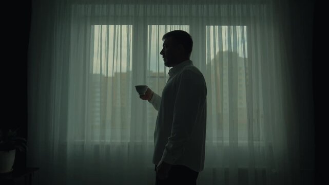 Morning coffee at the beginning of a hard day. Silhouette of a man in a shirt drinking tea from a mug against the background of a window, symmetrical cinematic frame.
