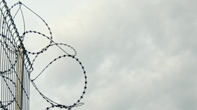 Close-up video of razor wire mounted on a tall metal security fence against an overcast sky. Symbolic security, restricted access, border protection, prison perimeter, surveillance, safety.