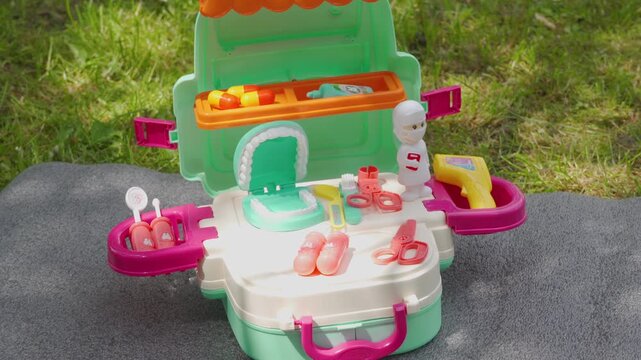 Colorful plastic dentist playset in the park. Toy doctor kit suitcase opened on a picnic blanket outdoors.