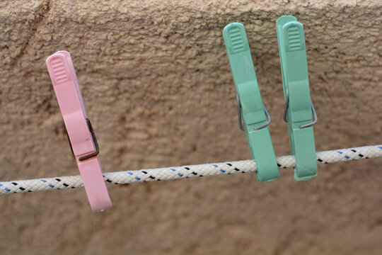 Three plastic clothespins in pink and light green colors are clipped onto a white braided clothesline against a textured concrete wall.