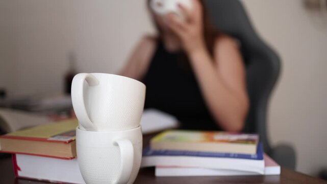 Coffee cups pile up on the desk as a young woman reaches for another refill hoping caffeine will restore focus during long hours of studying and mental fatigue
