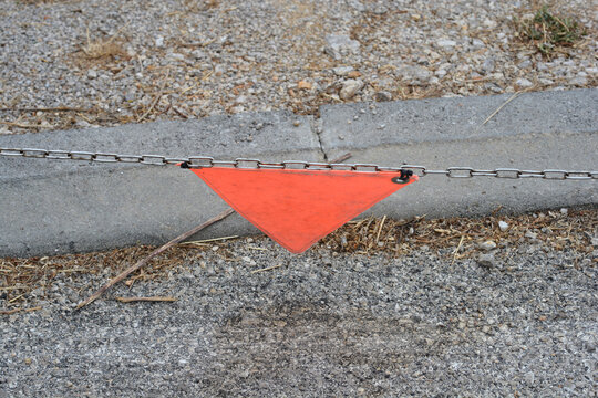 An orange plastic warning triangle is attached to a horizontal metal chain barrier above a concrete curb and asphalt ground.