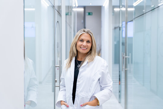 Woman medical professional wearing white coat smiling, standing in a clinic hallway