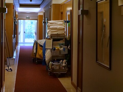 A long indoor hotel corridor. housekeeping cart stacked with white linens and cleaning supplies. Light streams in from glass exit doors at the end 
