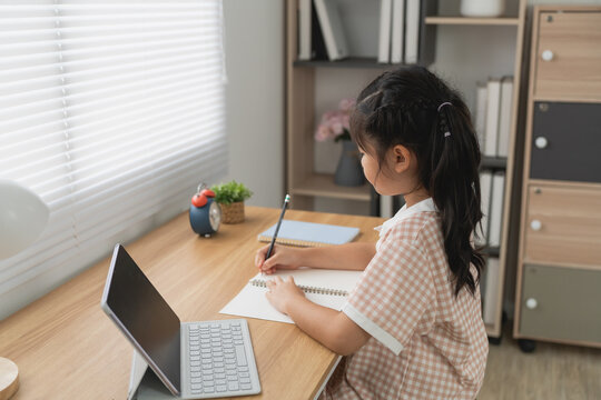 Young girl studying at wooden desk using tablet and notebook in modern bright room with natural light blinds