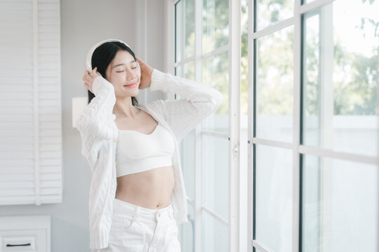 Young woman in a white sweater with headphones around her neck smiling by a bright window at home, enjoying a calm morning and fresh natural light.