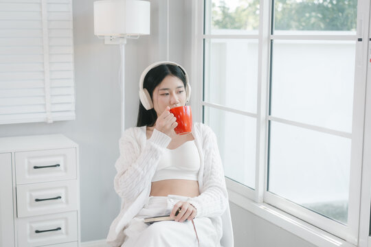 Young woman in white loungewear sits by a bright window, wearing headphones and drinking from a red mug in a calm modern home setting