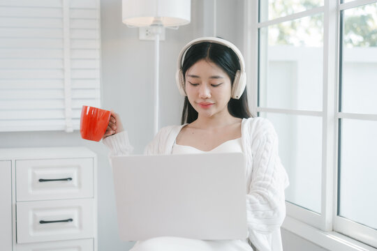 Young woman in white loungewear sits by a bright window, wearing headphones and drinking from a red mug in a calm modern home setting