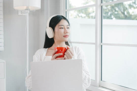 Young woman in white loungewear sits by a bright window, wearing headphones and drinking from a red mug in a calm modern home setting