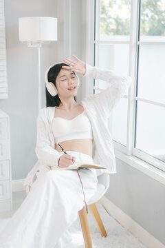 Young woman in white casual clothes using a smartphone and wearing headphones by a bright window at home, enjoying music and a relaxed morning mood.