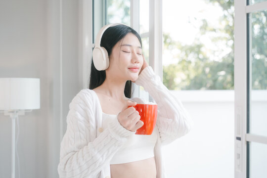 Young woman in white loungewear sits by a bright window, wearing headphones and drinking from a red mug in a calm modern home setting