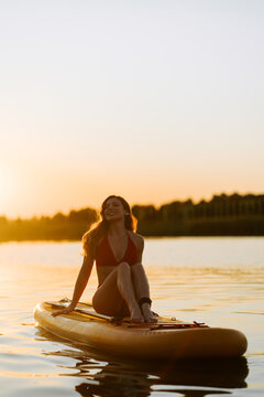 Young woman sitting on a paddle board in the middle of a calm lake during a bright golden sunset