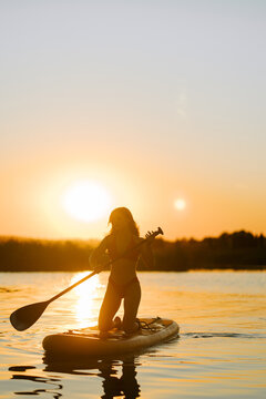 Woman kneeling on a paddle board while paddling across a quiet lake during a beautiful golden sunset
