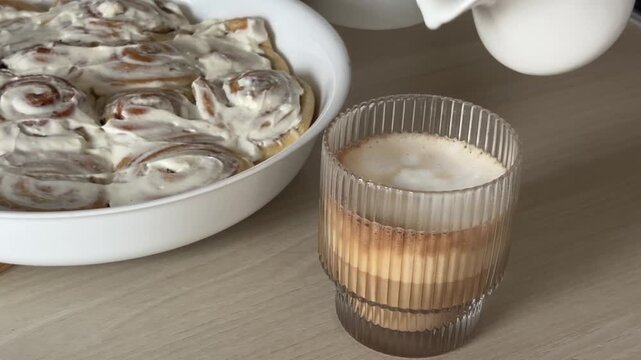 Pouring smooth velvety milk foam into a cup of coffee with freshly glazed cinnamon cream rolls lying on a side plate