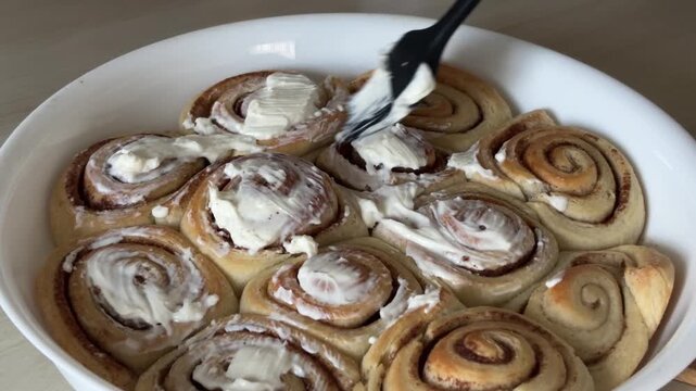 Spreading rich white buttercream frosting over hot fresh cinnamon rolls in a baking dish, sweet dessert preparation