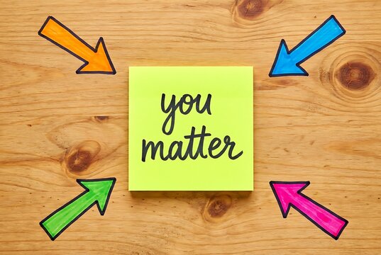 Bright yellow sticky note with text you matter written in black ink surrounded by colorful arrows pointing towards center on wooden background.