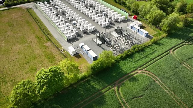 Aerial footage of a utility-scale battery energy storage system BESS with rows of battery containers and electrical substation next to green fields, camera pull out and tilt up.