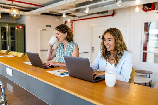 Two female professionals using laptops, drinking coffee, and collaborating in a modern coworking office for business and productivity