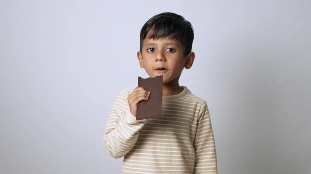 Indian boy eating chocolate bar. Happy child enjoying sweet snack on white background.