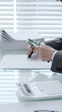 Hand holding magnifying glass and pen, studying financial paperwork and reviewing an invoice with a calculator on desk, performing a detailed audit and verification of data