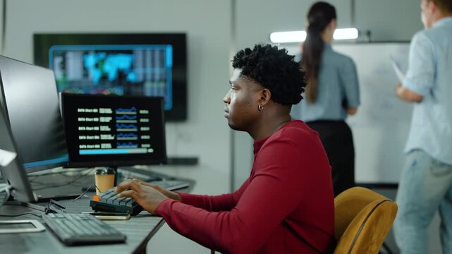 Focused Black male data analyst works on a multi-monitor desk in a busy corporate office. smiling female Asian colleague approaches, points at the computer screen, and they collaborate on a project