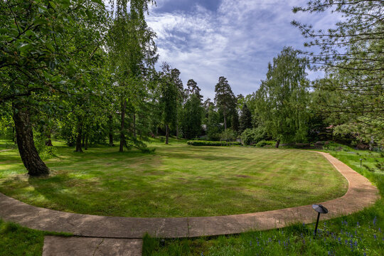 A park is seen with lush green grass and tall trees. A curved path leads through the area. It is bright and sunny, ideal for a walk or outdoor activity