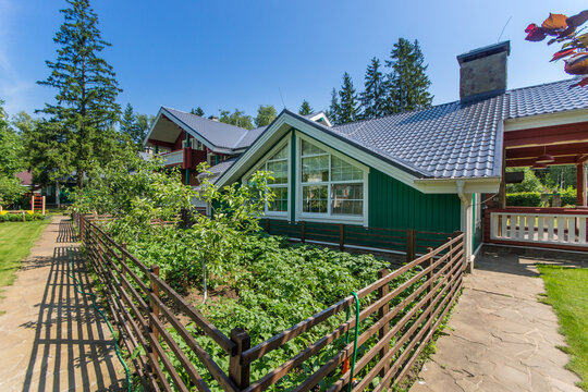 A green house stands beside a well-kept garden surrounded by a wooden fence. Trees provide shade in the background under a bright blue sky. The scene shows clear weather and nature.