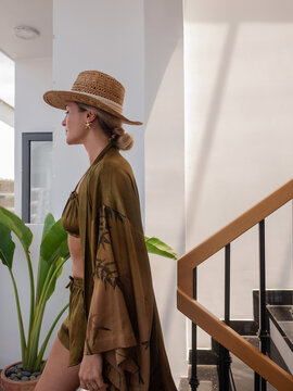 Woman in olive resort wear and straw hat posing by modern staircase with tropical plant and natural sunlight