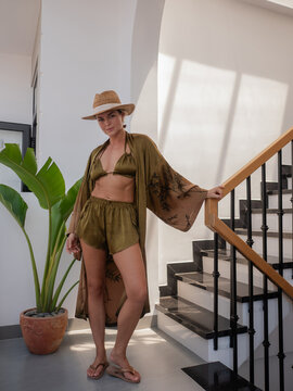 Woman in olive resort wear and straw hat posing by modern staircase with tropical plant and natural sunlight