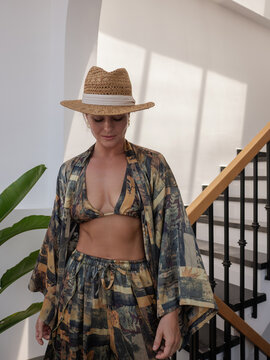 Woman modeling patterned resort wear with bikini top, flowing robe and straw hat in sunlit modern interior