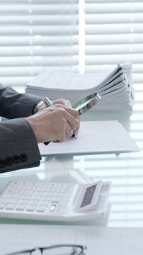Hand holding magnifying glass and pen, studying financial paperwork and reviewing an invoice with a calculator on desk, performing a detailed audit and verification of data