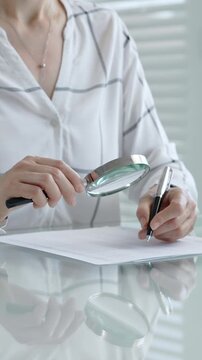 Woman's hands using a magnifying glass to scrutinize a document while holding a pen, researching important information for legal, financial, or academic analysis and validation