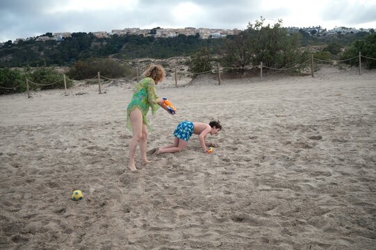 Mother playfully chasing her teenage son with water guns during a fun family day on a sandy beach