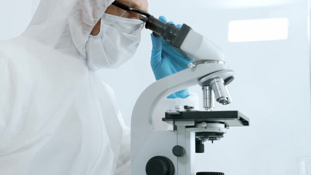 Scientist wearing a hazmat suit, face mask, protective glasses, and blue gloves, looking through a microscope in a clean room, performing critical research and analysis