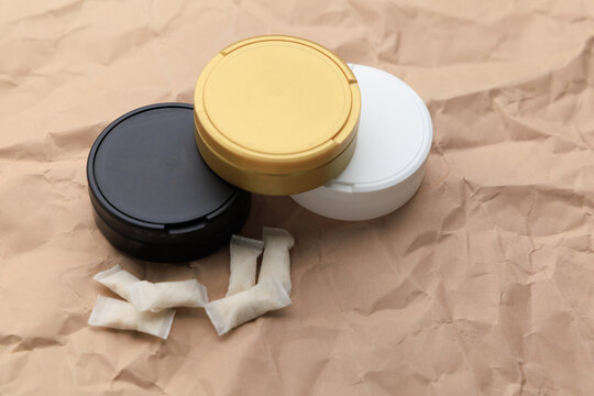 Different containers hold snus pouches and nicotine pads in a simple setting on brown textured paper