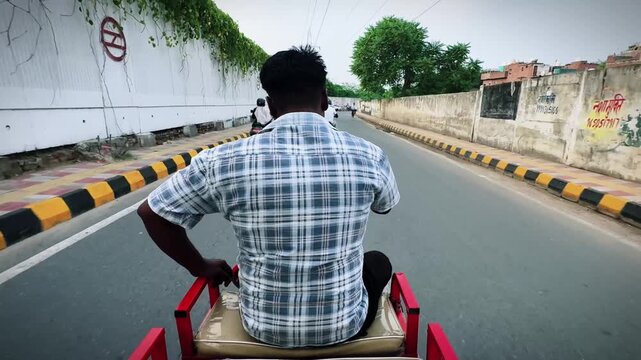 Rear view of an auto rickshaw rider driving along a quiet urban road during daylight. The image captures everyday transportation, city commuting, and the daily life of a rickshaw driver in India.