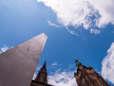 Prague Castle Monolith and St. Vitus Cathedral seen from a dramatic low angle in Prague, Czechia. Monumental granite form and Gothic towers rise against a vivid blue sky and white clouds.
