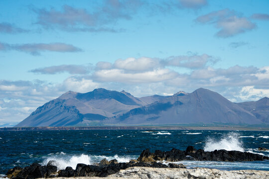 Ocean waves crashing on the rocky shore with beautiful mountains under blue sky in Iceland.
