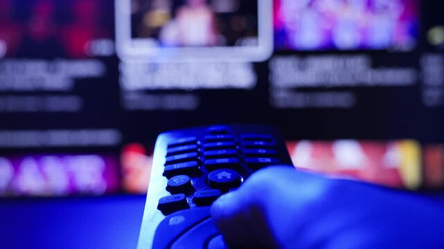 Close-up of hand holding TV remote control and choosing streaming content on smart TV at home. Watching movies and digital entertainment concept. High quality 4k footage
