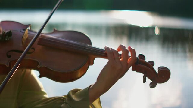 Close-up of a violinist playing soothing classical music outdoors by a tranquil lakeside at sunset, highlighting delicate finger placement and natural reflections