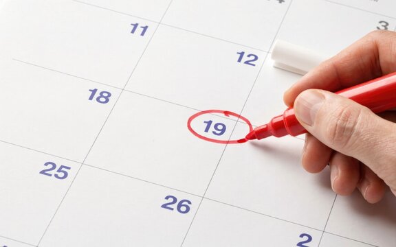 Hand holding a vibrant red marker circles the number nineteen on a white paper calendar to signify an important meeting date or a special deadline for a busy professional schedule.