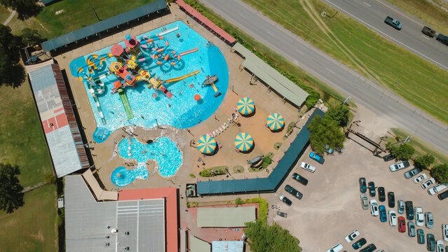 Interstate 35W lies adjacent to water park grounds near Fort Worth. Bird's eye view captures colorful splash fortress, swimming pools, umbrella shades under bright summer sunlight, Burleson, TX