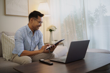Fototapeta premium Smiling businessman using a digital tablet and laptop while relaxing on a sofa in a modern living room.