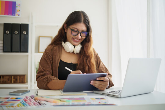 Smiling female graphic designer using a stylus and digital tablet while working with a laptop in a bright home office.