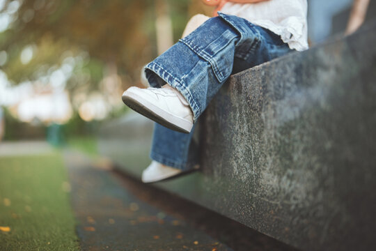 Close up legs of Asian toddler wearing denim jeans and white sneakers swinging feet while sitting on concrete ledge at city park during sunset, little child leisure fashion and outdoor activity