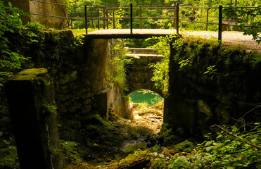 Obraz premium Mossy stone sluice structure and footbridge overlooking emerald green river water in a dense summer forest near Idrija, Slovenia.