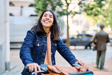 Fototapeta premium A young woman walks her bike along a lively city street, smiling and enjoying an autumn day. She wears a denim jacket and backpack, suggesting a casual commute or urban errand. The relaxed mood and na