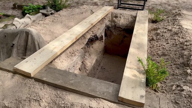 Freshly excavated open grave prepared for burial in a peaceful cemetery in Latvia with wooden support planks, sandy soil and funeral setup before coffin arrival