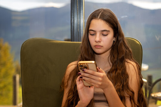 Teenage Girl Using Smartphone by a Mountain View Window. Teenage girl seated by a window using a smartphone with a blurred mountain landscape in the background.
