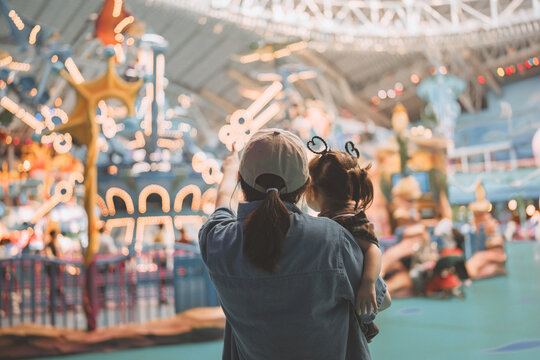 Back view of Asian mother holding toddler daughter pointing at carousel lights in indoor amusement park, happy family enjoying theme park vacation for childhood experience, entertainment and travel co